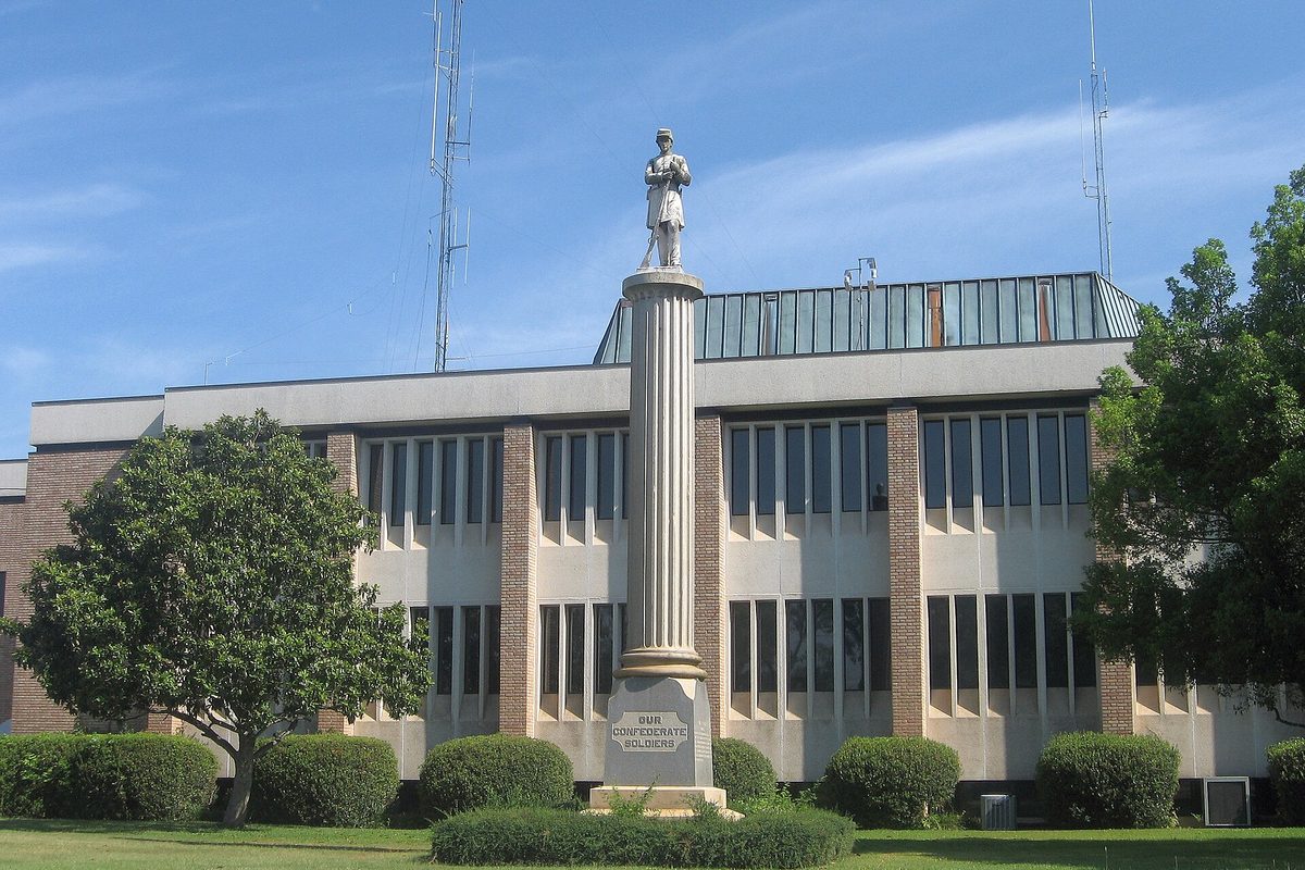 The Greenwood County Courthouse, a modern building with tall windows, behind a stone column topped by a soldier statue and a magnolia tree