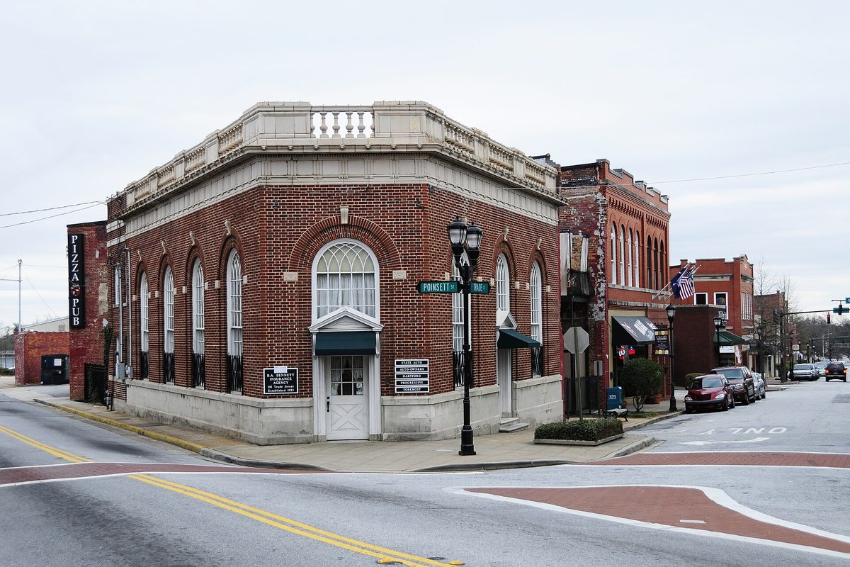 A red brick corner building with tall arched windows and a stone balustrade on a downtown Greer street, with a Poinsett Street sign and more brick storefronts behind
