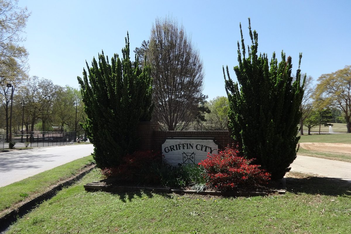 A Griffin City sign in a brick planter with red azaleas, between two tall evergreen shrubs at the edge of City Park