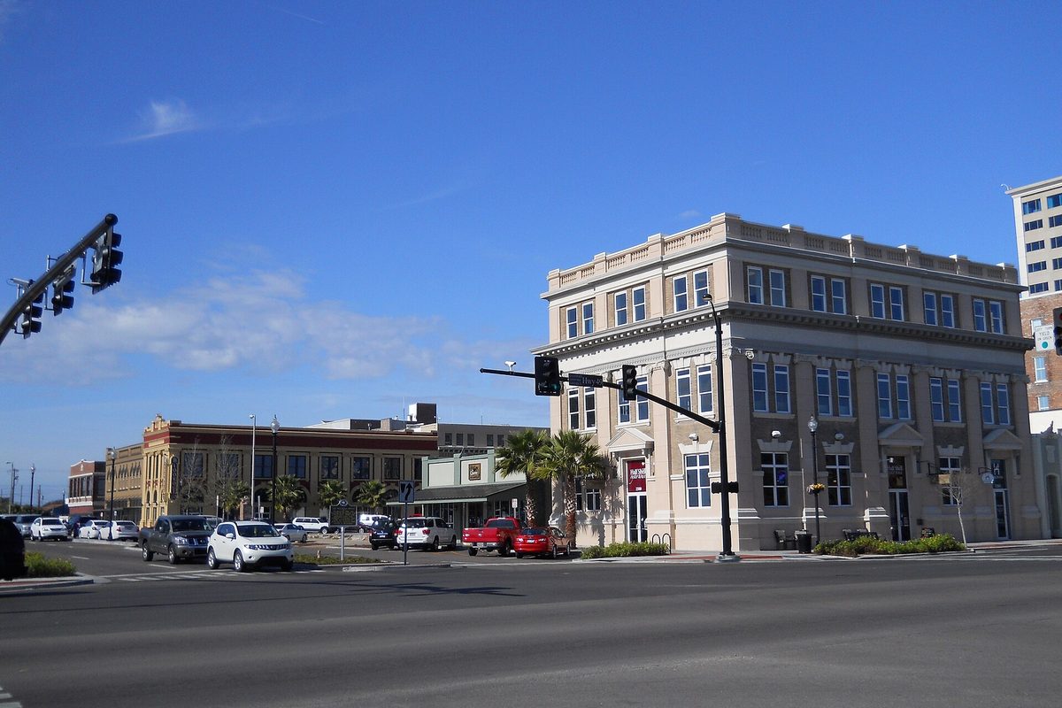 A cream three-story corner building and older brick storefronts at 25th Avenue and 13th Street in downtown Gulfport under a blue sky