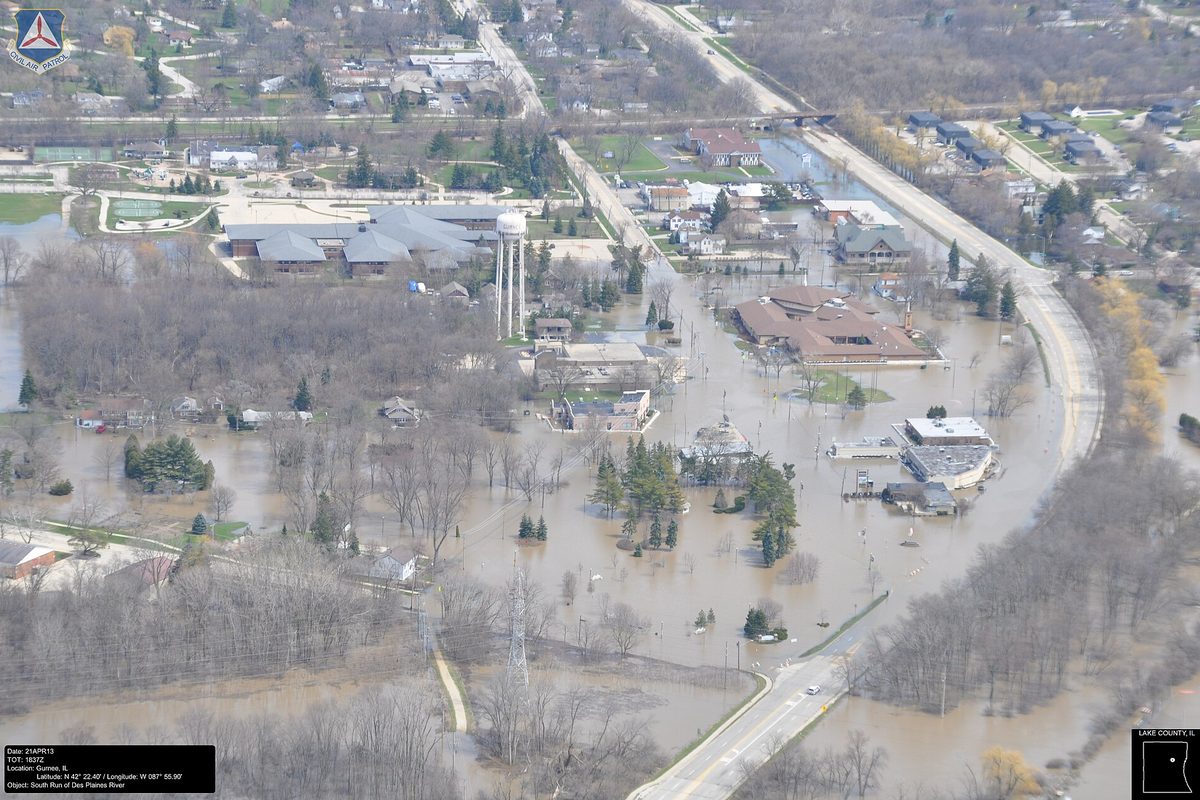 Aerial view of the Des Plaines River over its banks in Gurnee, with brown floodwater around buildings, roads and a white water tower