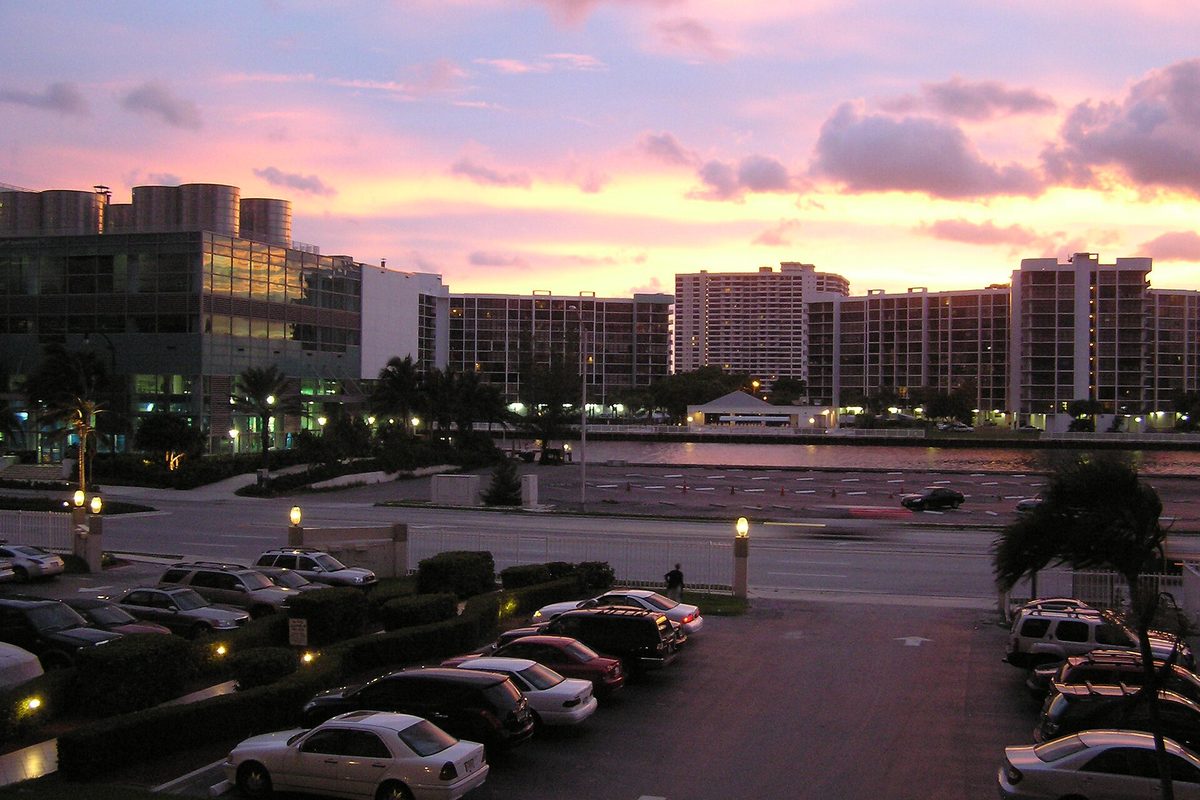 Pink and orange sunset over Hallandale Beach high-rises, with a lit parking lot full of cars in front and a waterway behind