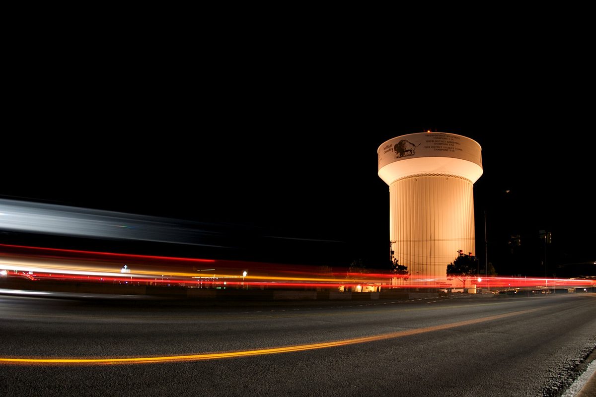 A Haltom City water tower lit up at night beside a road, with red and white light streaks from passing cars