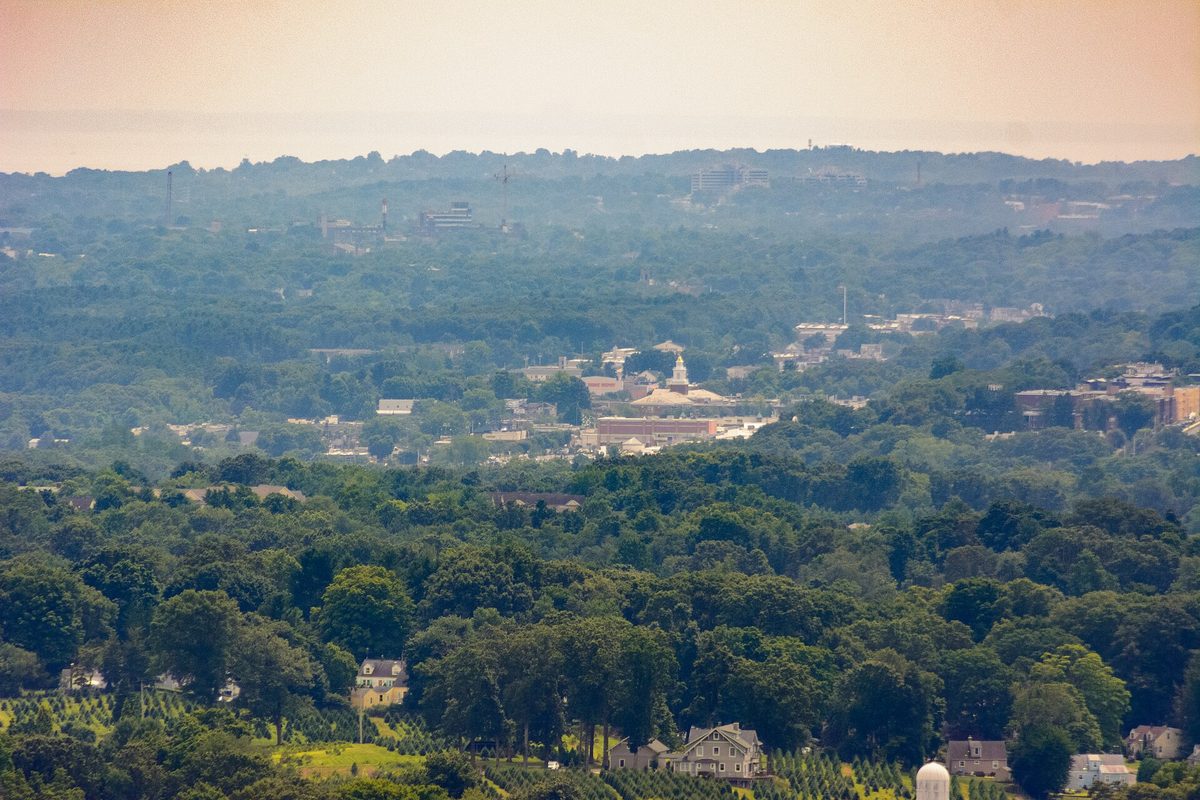 Hazy view from the White Trail at Sleeping Giant over the green treetops of Hamden, with the buildings of Hamden High School in the middle distance and low hills beyond