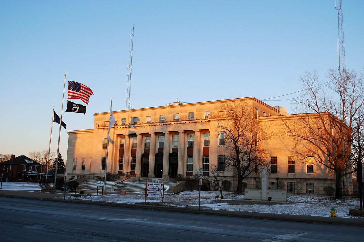 Hammond City Hall, a pale stone building with tall columns, flags flying out front and a thin layer of snow on the lawn at sunrise