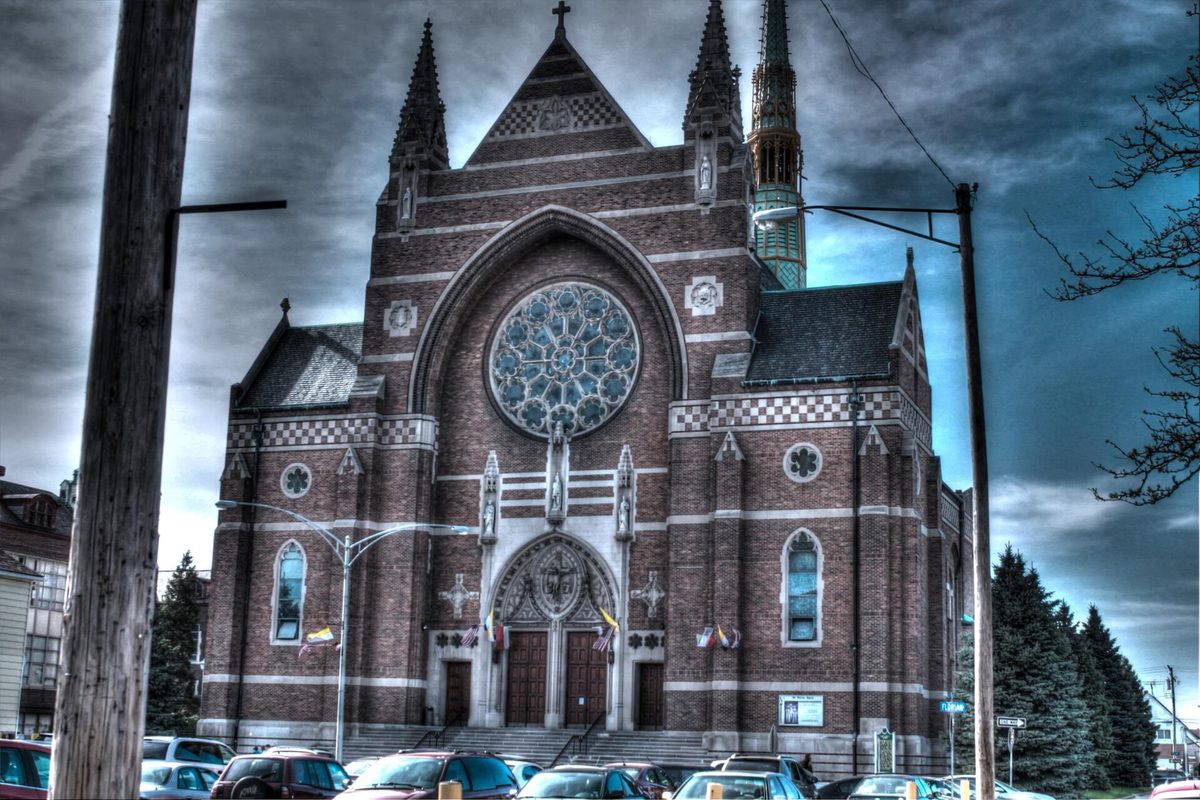 The red brick St. Florian church in Hamtramck, with a large rose window and a green-capped spire, behind a row of parked cars under a dark sky