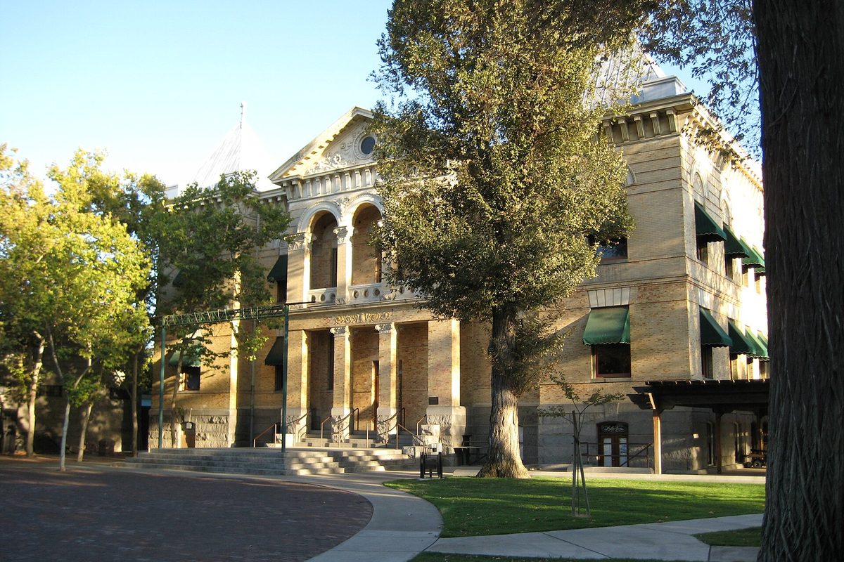 The tan brick Kings County Courthouse in Hanford with arched windows, green awnings and a large tree on the lawn in front