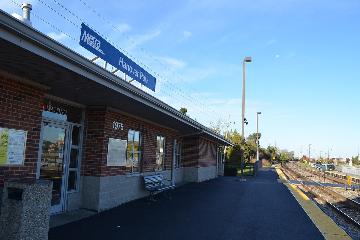 The brick Hanover Park Metra station with a blue Metra sign, a bench by the waiting room door and an empty platform beside the tracks