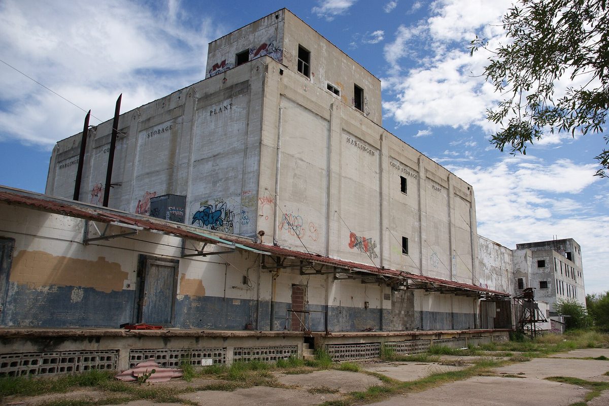 The abandoned Harlingen Cold Storage building, a tall concrete warehouse with broken windows, graffiti and a rusted loading-dock awning above a cracked, weedy lot