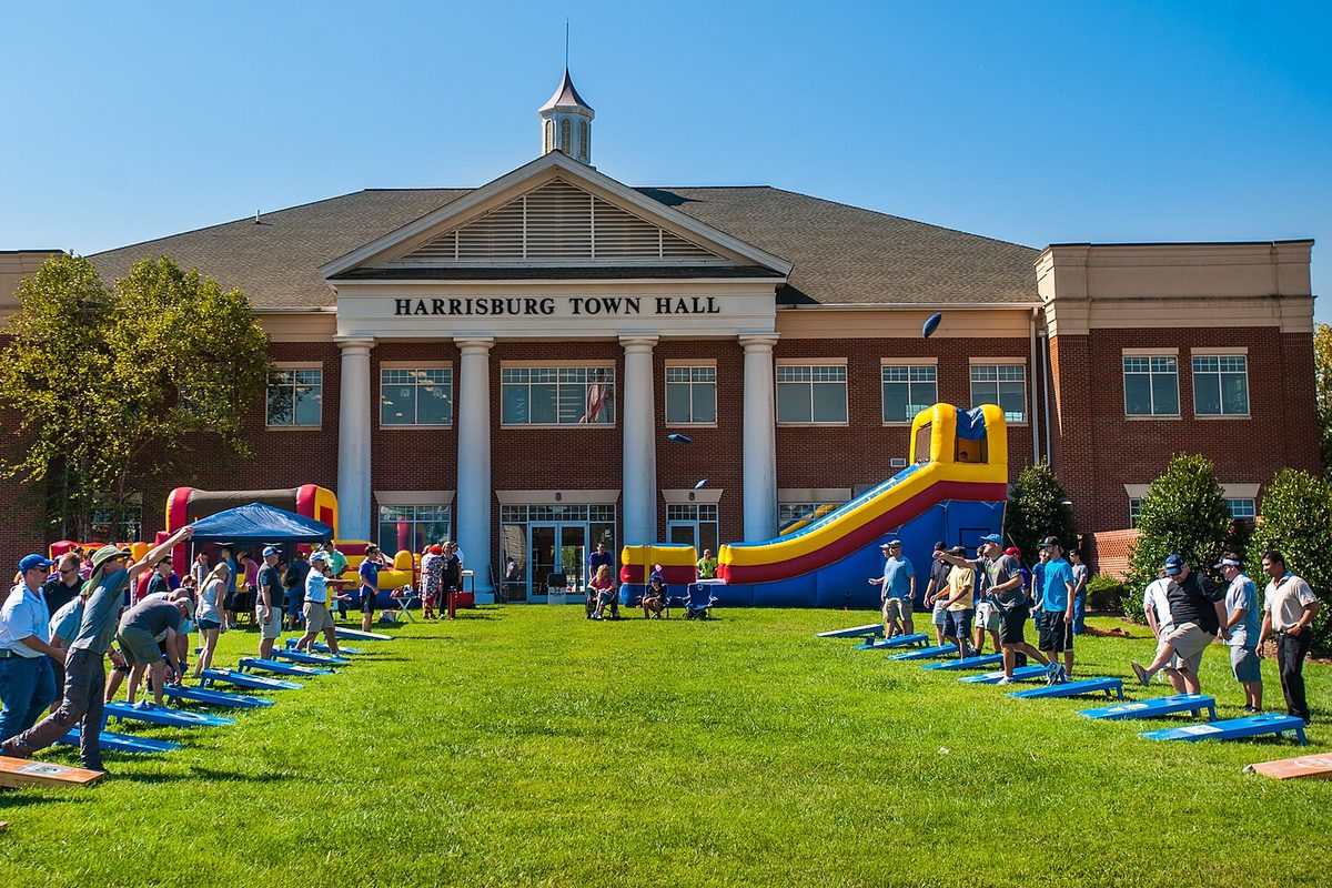 Harrisburg Town Hall, a brick building with white columns and a cupola, with people playing cornhole and an inflatable slide on the front lawn