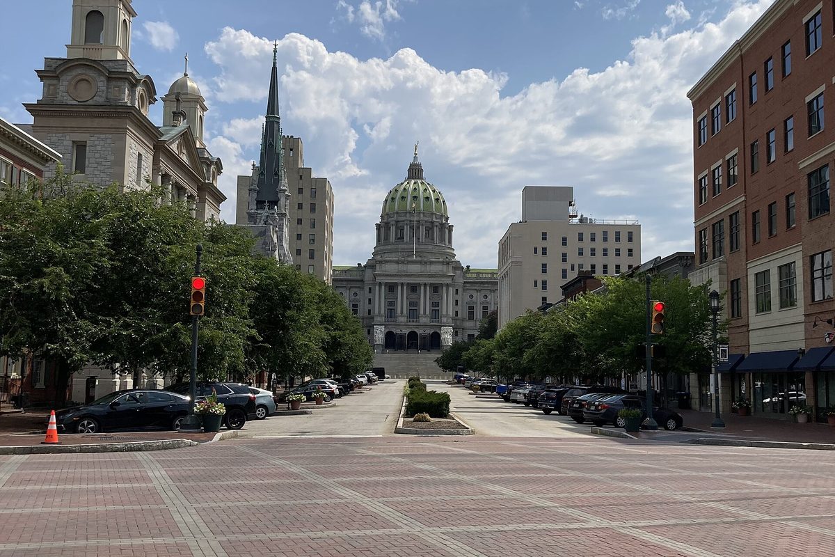 The green dome of the Pennsylvania State Capitol at the end of a brick-paved downtown Harrisburg street, with church steeples, trees and parked cars on both sides