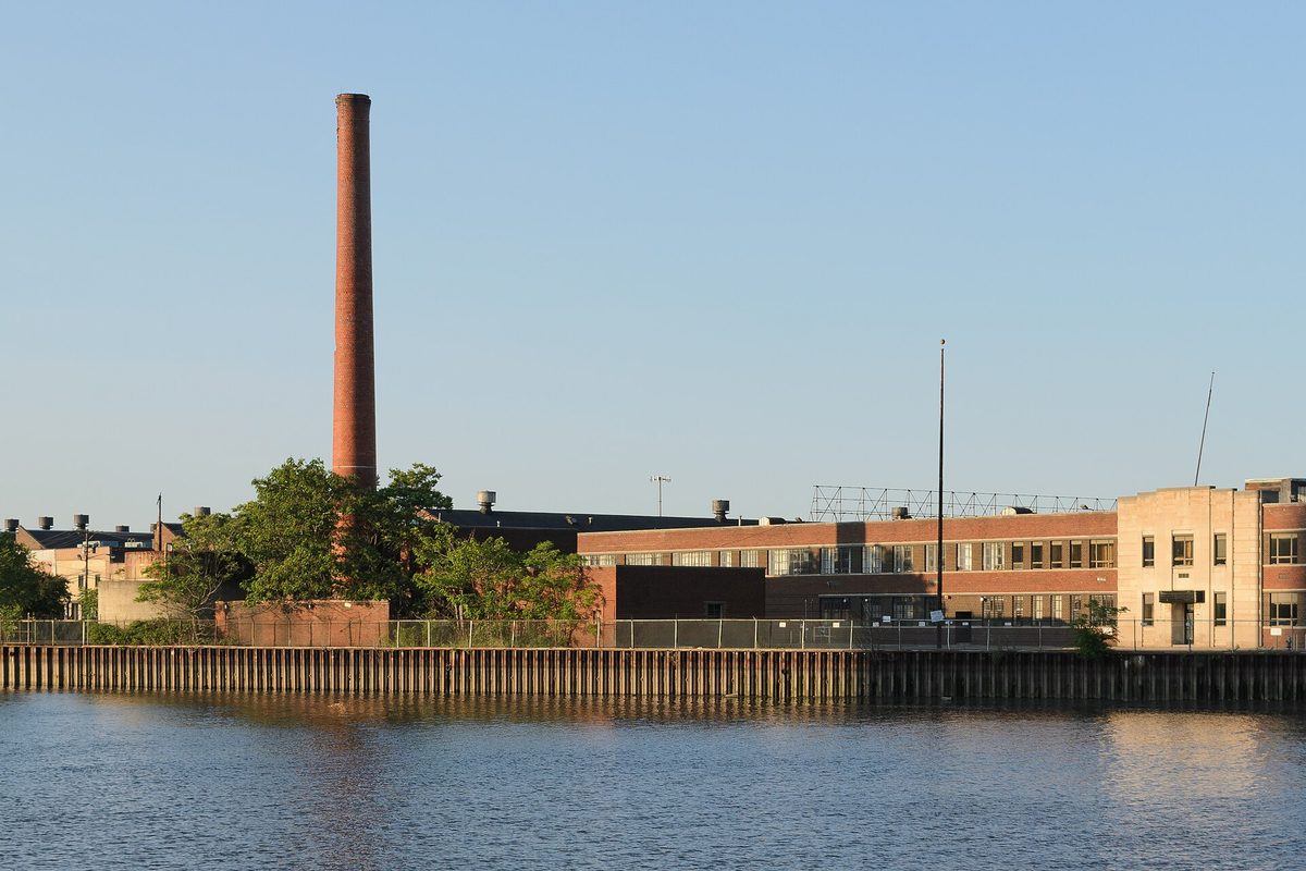 Brick industrial buildings and a tall brick smokestack along the river in Harrison, seen across the water in early evening light