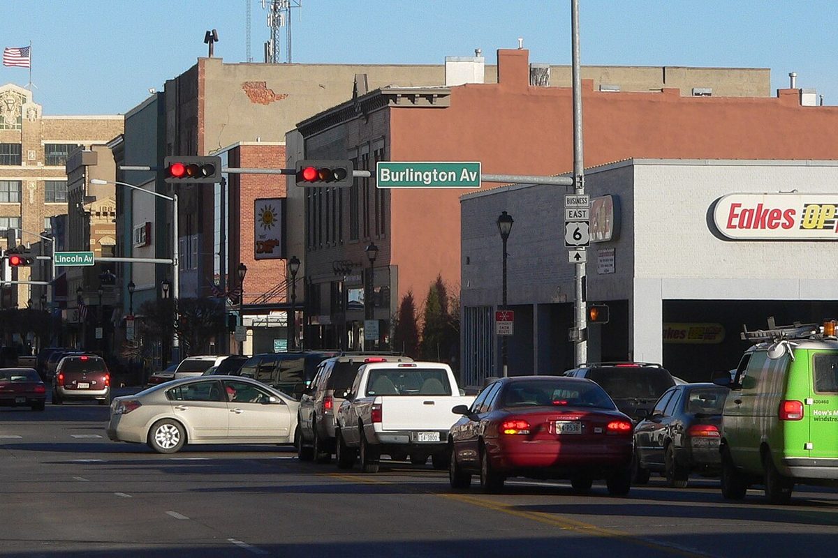 Traffic stopped at a red light at Burlington Avenue in downtown Hastings, with brick buildings, a white pickup and a red sedan in the lanes and a US 6 East sign on the signal pole