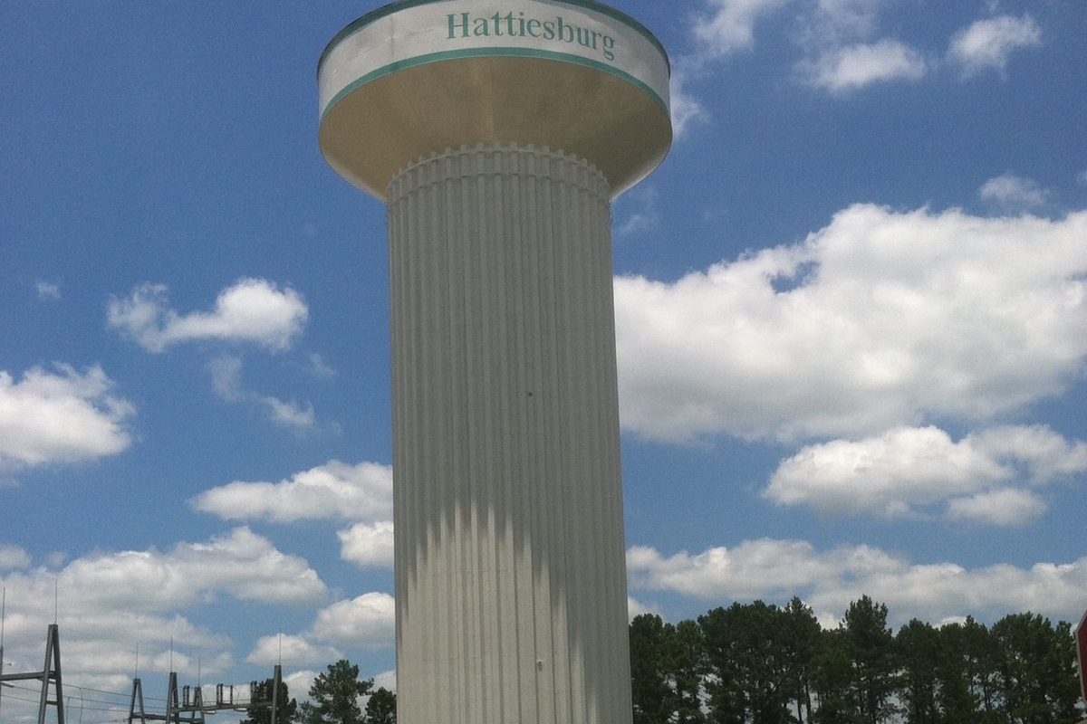 A tall white water tower with Hattiesburg lettered on its tank, against a blue sky with white clouds