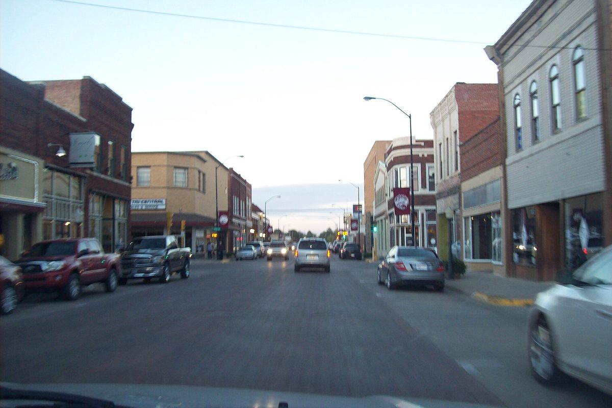 Main Street in downtown Hays at dusk, seen through a car windshield, with brick storefronts, street lamps and cars along both curbs