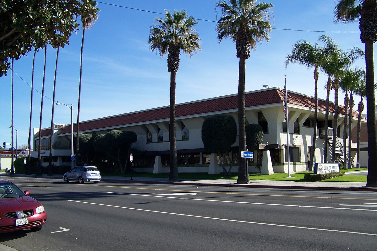 Hemet City Hall, a low white building with a red tile roof and tall palm trees, beside a street with a red car parked at the curb