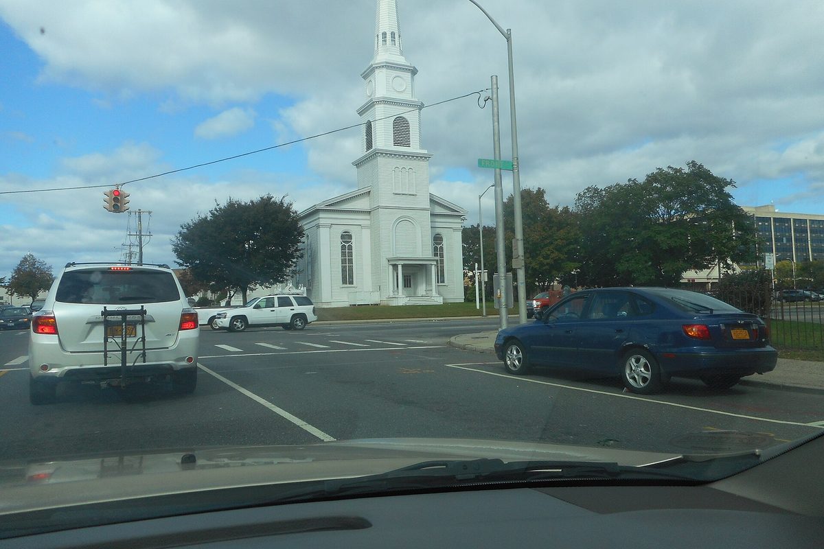 The white United Methodist Church of Hempstead with its tall steeple at a street corner, seen through a car windshield with traffic at the light