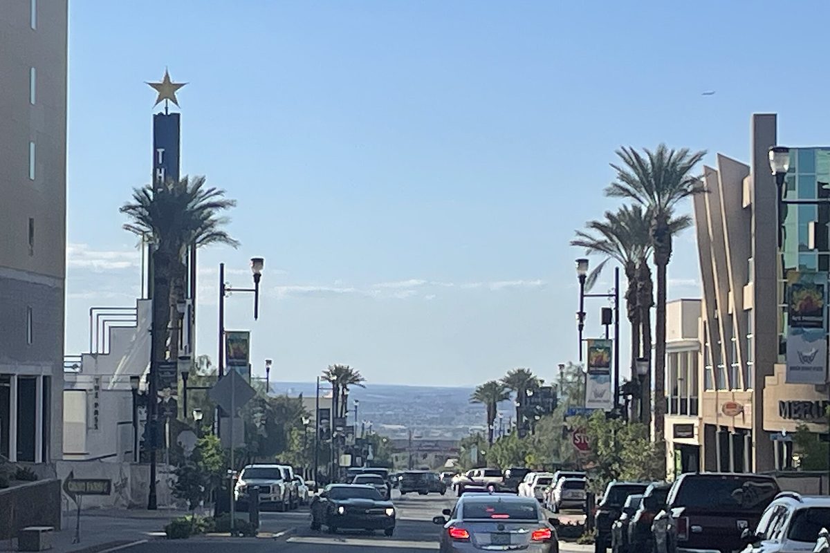 A street in Henderson's Water Street District lined with palm trees, with traffic, a tall sign tower topped by a gold star and hills in the distance