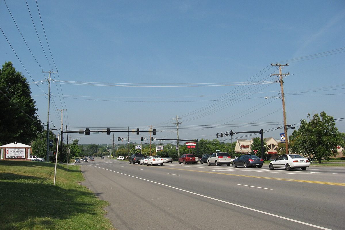 A wide multi-lane road through Hendersonville lined with utility poles, traffic signals and roadside business signs on a clear summer day