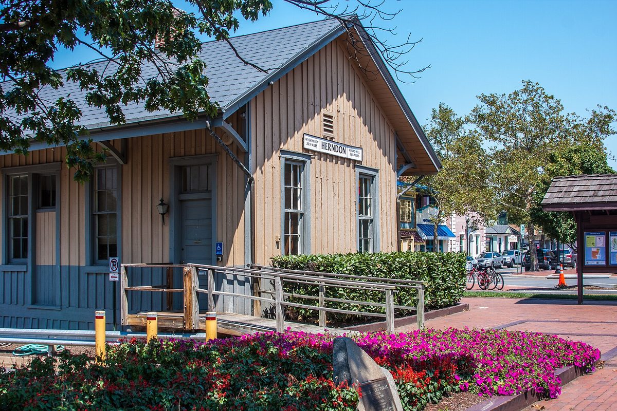The tan wooden Herndon Depot with its HERNDON sign, beds of pink and red flowers in front, and bicycles parked by a brick plaza on a sunny day