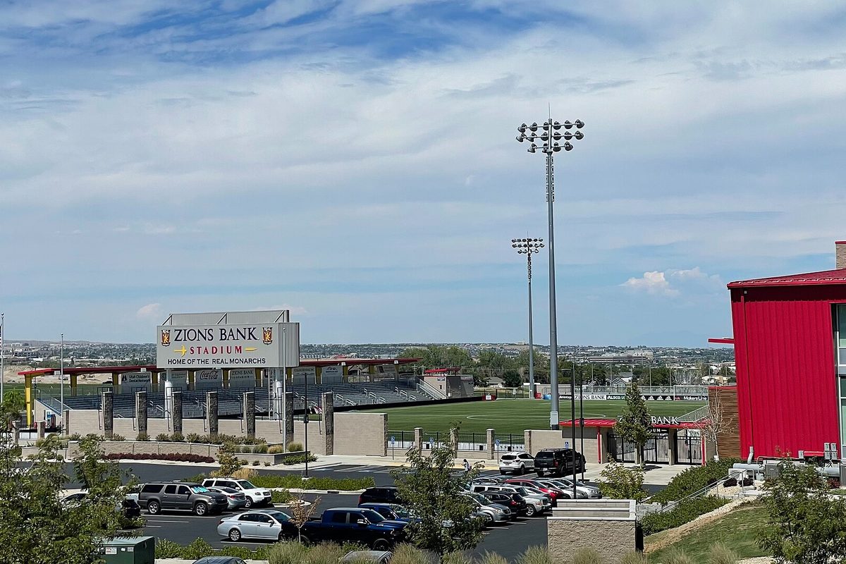 Zions Bank Stadium in Herriman, with its scoreboard sign, tall light towers and a parking lot of cars and pickups under a partly cloudy sky