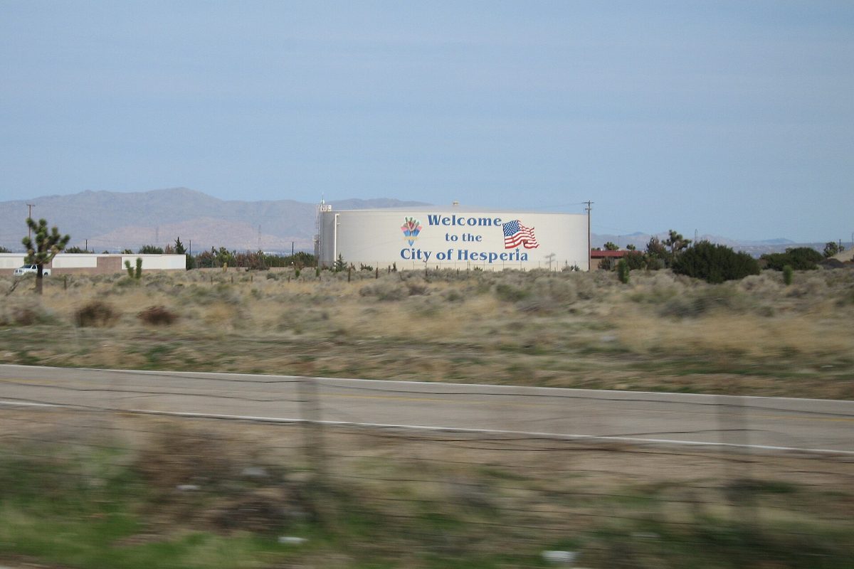 A white water tank painted Welcome to the City of Hesperia with an American flag, beside a desert road lined with Joshua trees and dry scrub