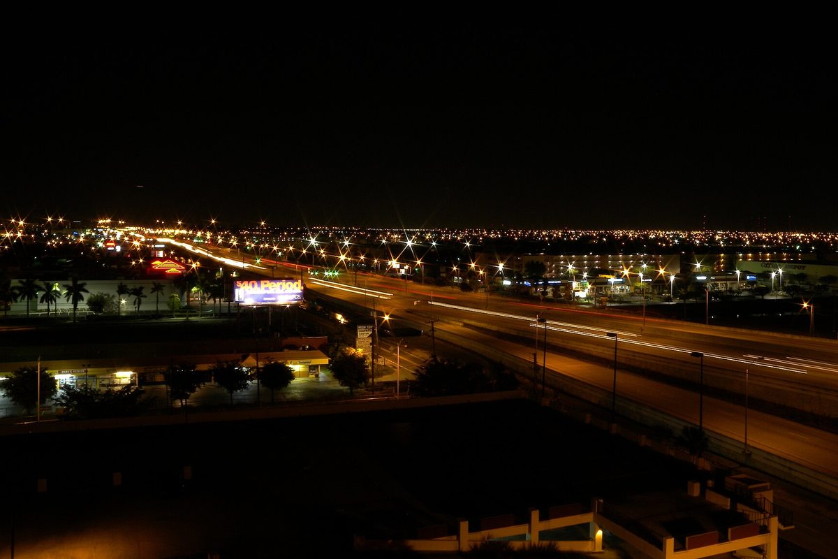 A divided highway at night seen from a height, with streaks of headlights, a lit billboard, palm trees and city lights running to the horizon