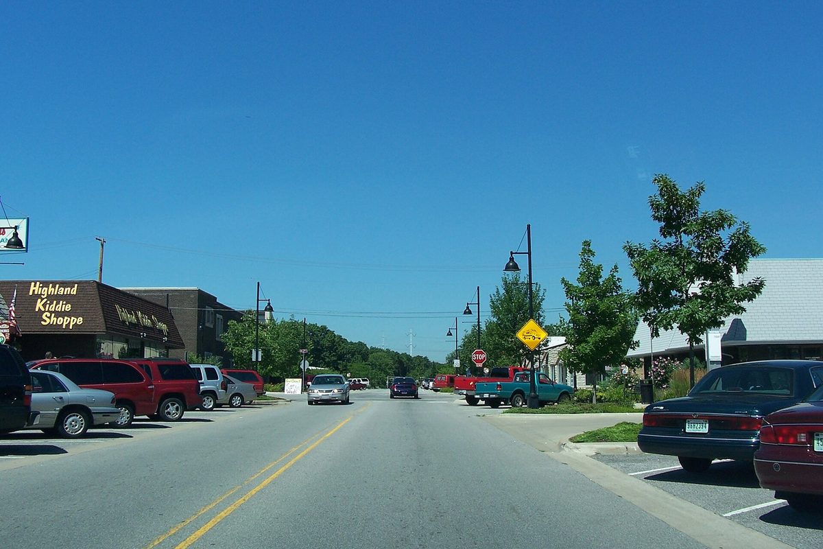 A street of low shops in Highland, Indiana, with the Highland Kiddie Shoppe sign, parked cars and pickups, lamp posts and young trees under a clear sky