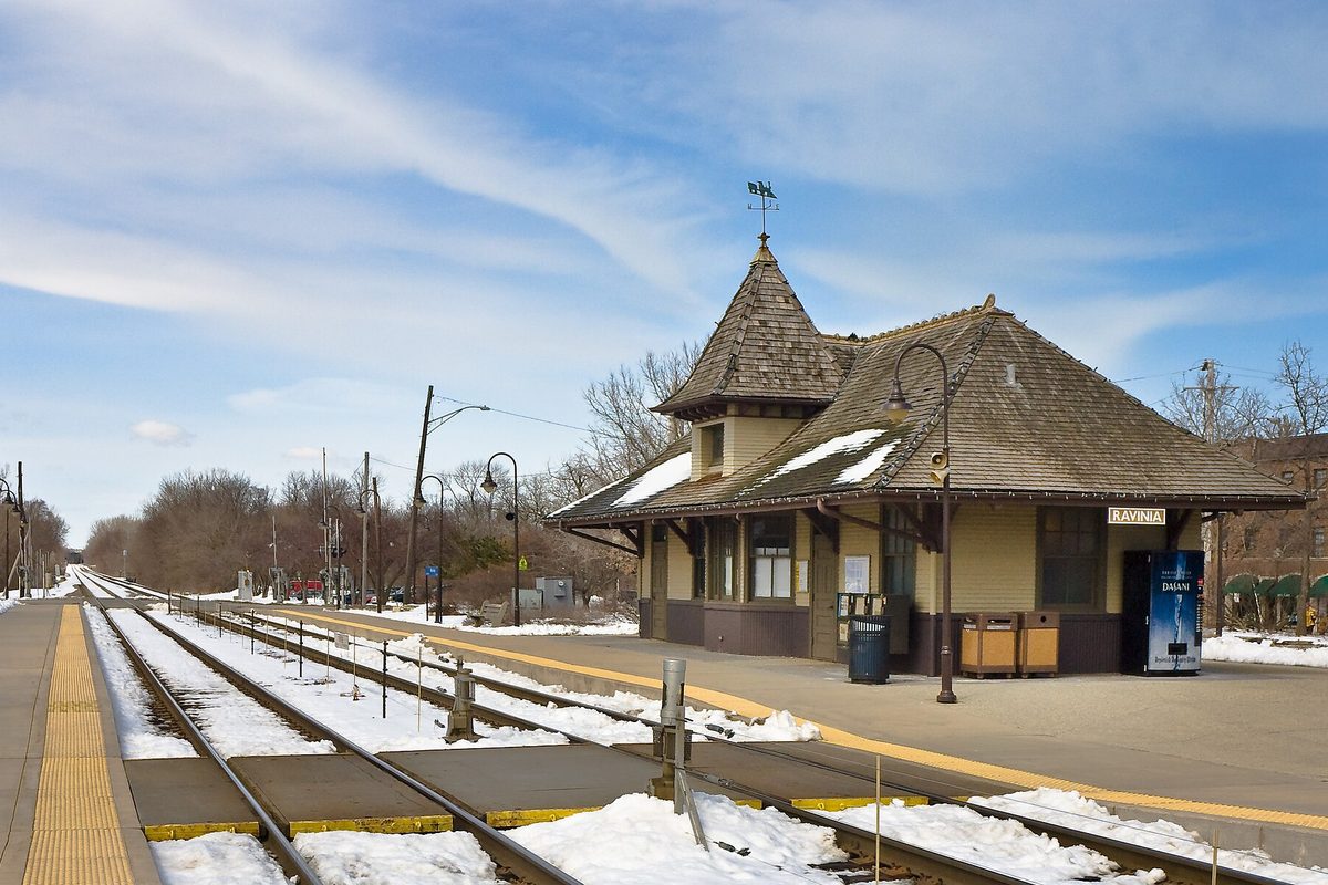 The brown shingled Ravinia Metra station in Highland Park, with a pointed roof and weather vane, beside snow-covered tracks under a blue sky