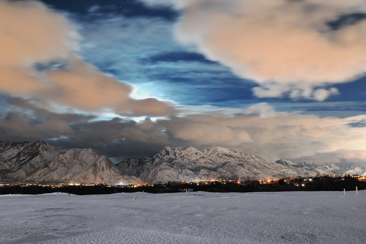 Highland at night under moonlit clouds, with snow-covered mountains above a band of town lights and a snowy field in the foreground