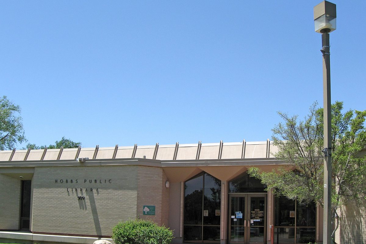 The low tan brick Hobbs Public Library with its name on the wall, glass entry doors and a tall street lamp under a clear blue sky