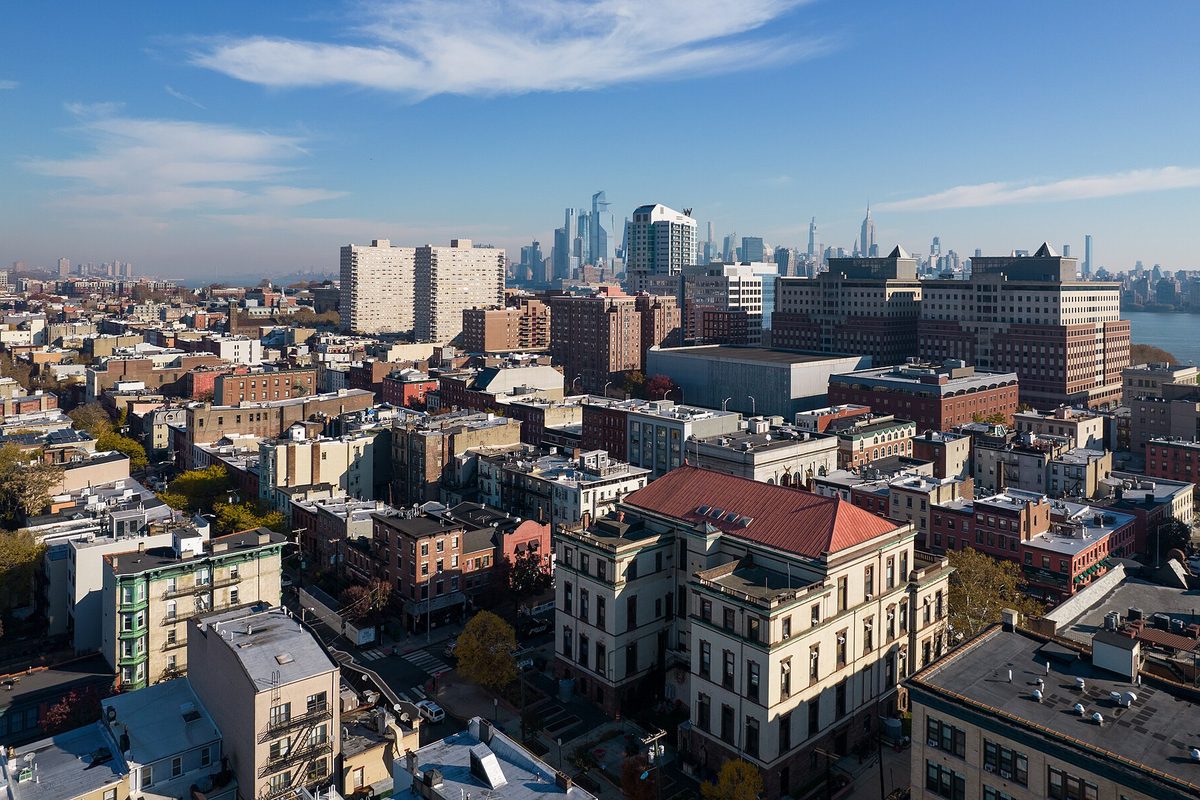 Aerial view to the northeast over Hoboken's brick rowhouses and apartment buildings, with the Manhattan skyline across the river under a blue sky