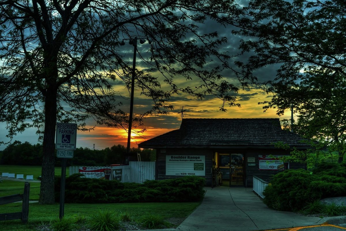 A small shingled building with a lit doorway behind shrubs and a parking sign, framed by tree silhouettes against an orange sunset in Hoffman Estates