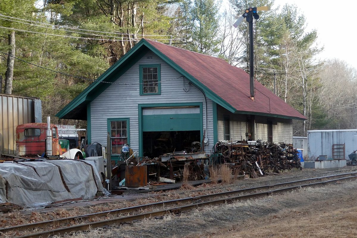 The former Holden rail station, a small gray building with a red roof and green trim beside the tracks, with scrap metal piled out front and an old rusted truck to the left