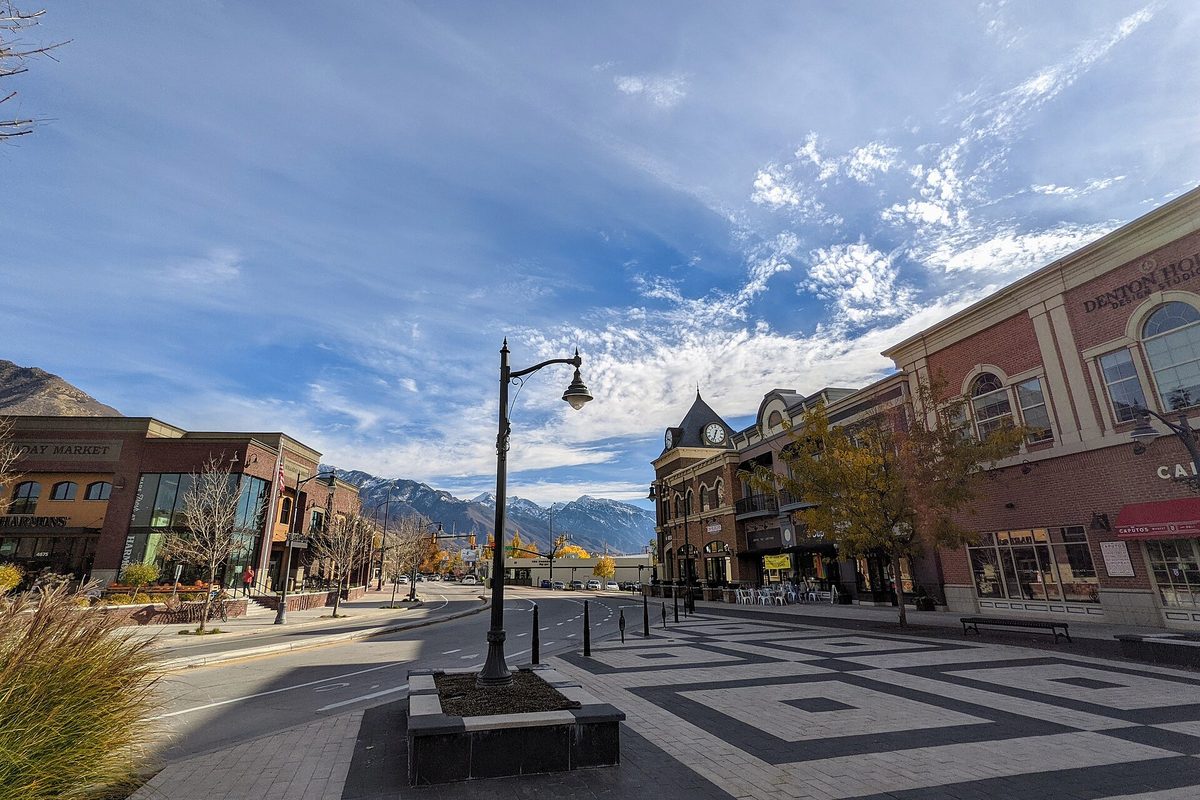 Brick shops and a clock tower around a paved plaza in Holladay Village, with autumn trees and snow-dusted mountains under a blue sky