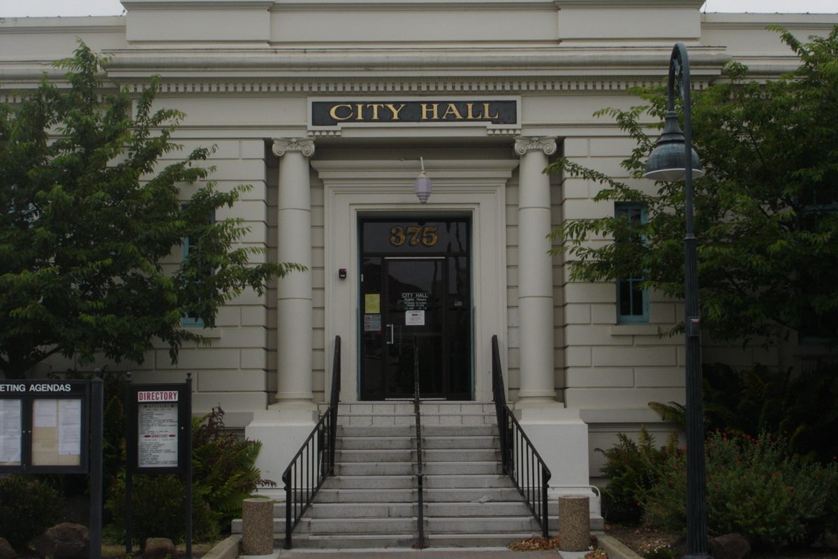 The columned stone entrance of Hollister City Hall, number 375, with steps, black handrails and a lamp post beside a tree