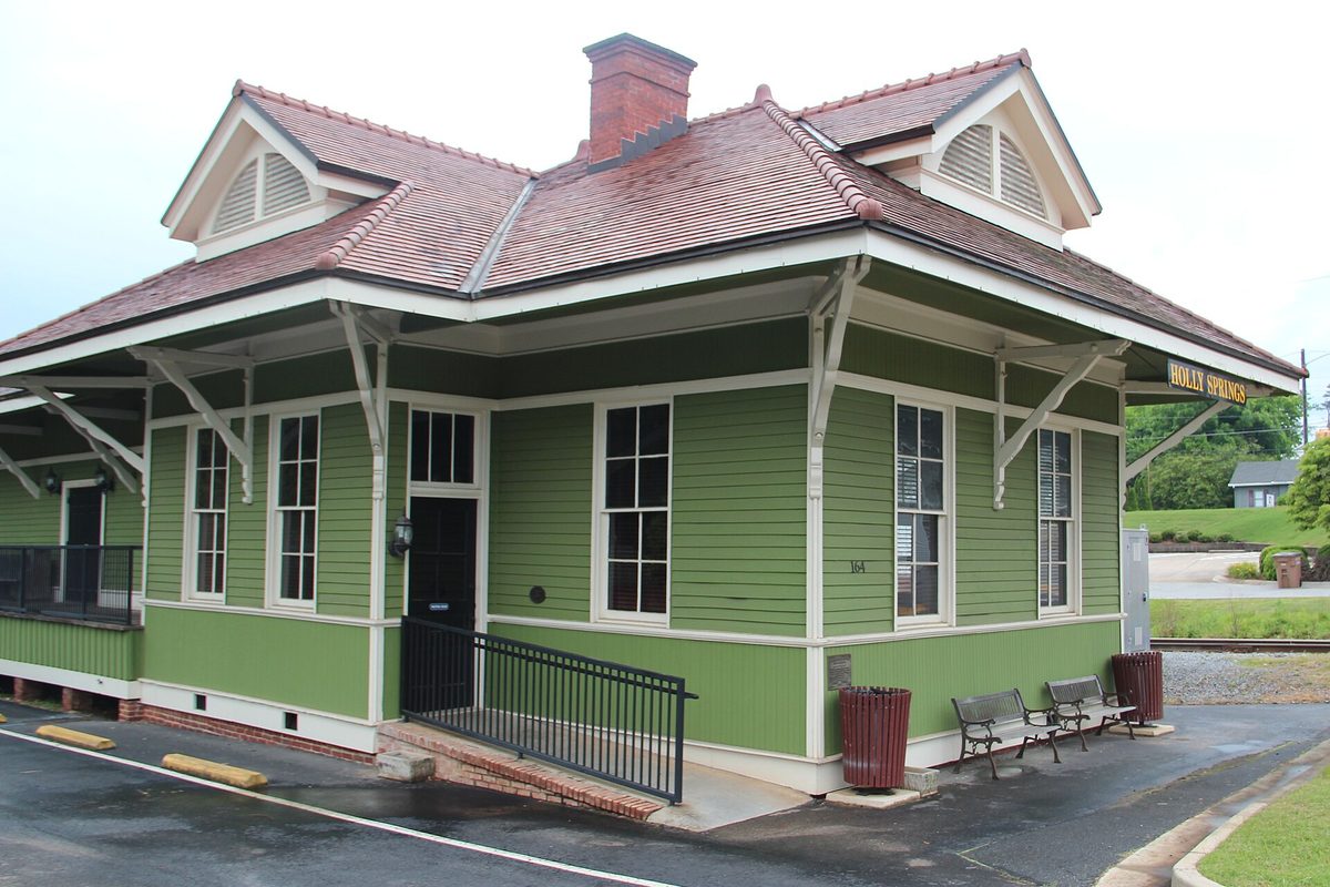 The restored Holly Springs train depot, a green wooden building with white trim, a red tile roof and a brick chimney, with benches along the platform