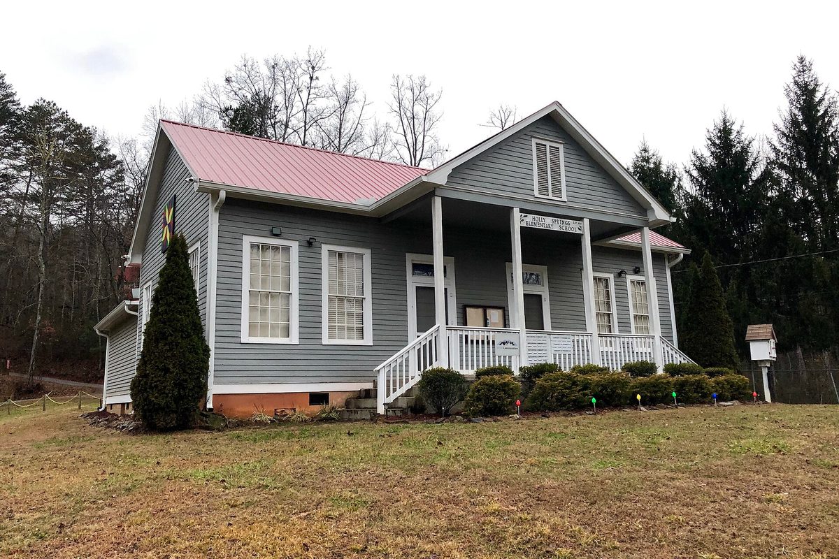 A gray clapboard former schoolhouse with a pink metal roof, white porch and steps, set on a lawn among bare trees and evergreens