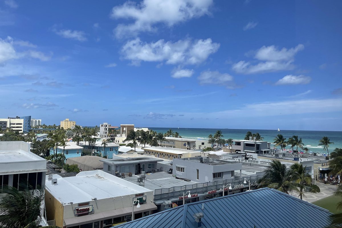 Low flat-roofed buildings and palm trees in Hollywood Beach, with the blue Atlantic behind them under a bright sky with scattered clouds