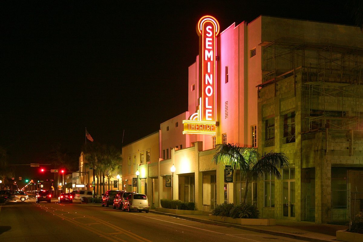 The Seminole Theatre's tall neon sign glowing red and orange at night above a quiet Homestead street with cars parked along the curb