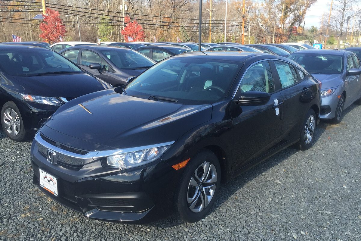 A black 2016 Honda Civic LX sedan on a gravel dealer lot, with more Civics and bare autumn trees behind it
