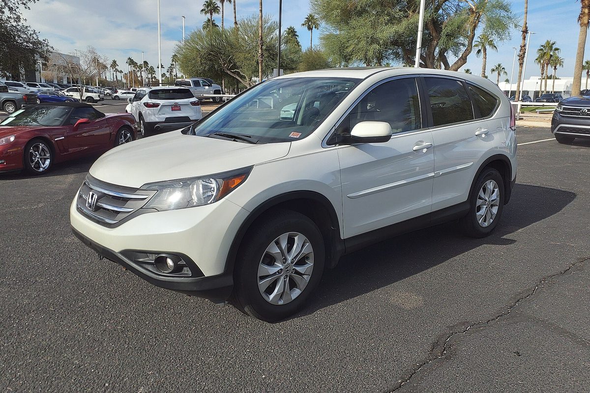 A white 2014 Honda CR-V EX parked on a sunny dealer lot, with palm trees and a red convertible behind it