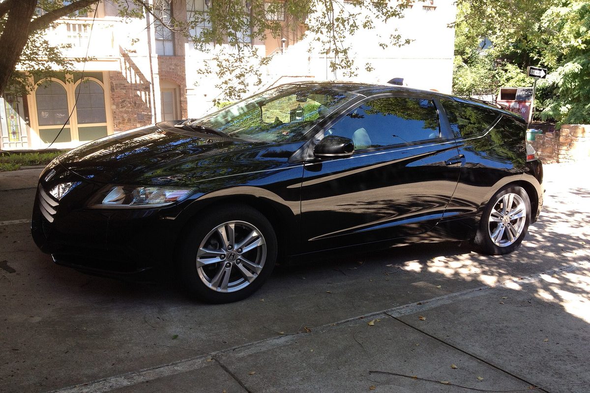 A black Honda CR-Z hybrid coupe parked in dappled shade on a concrete driveway in front of a house