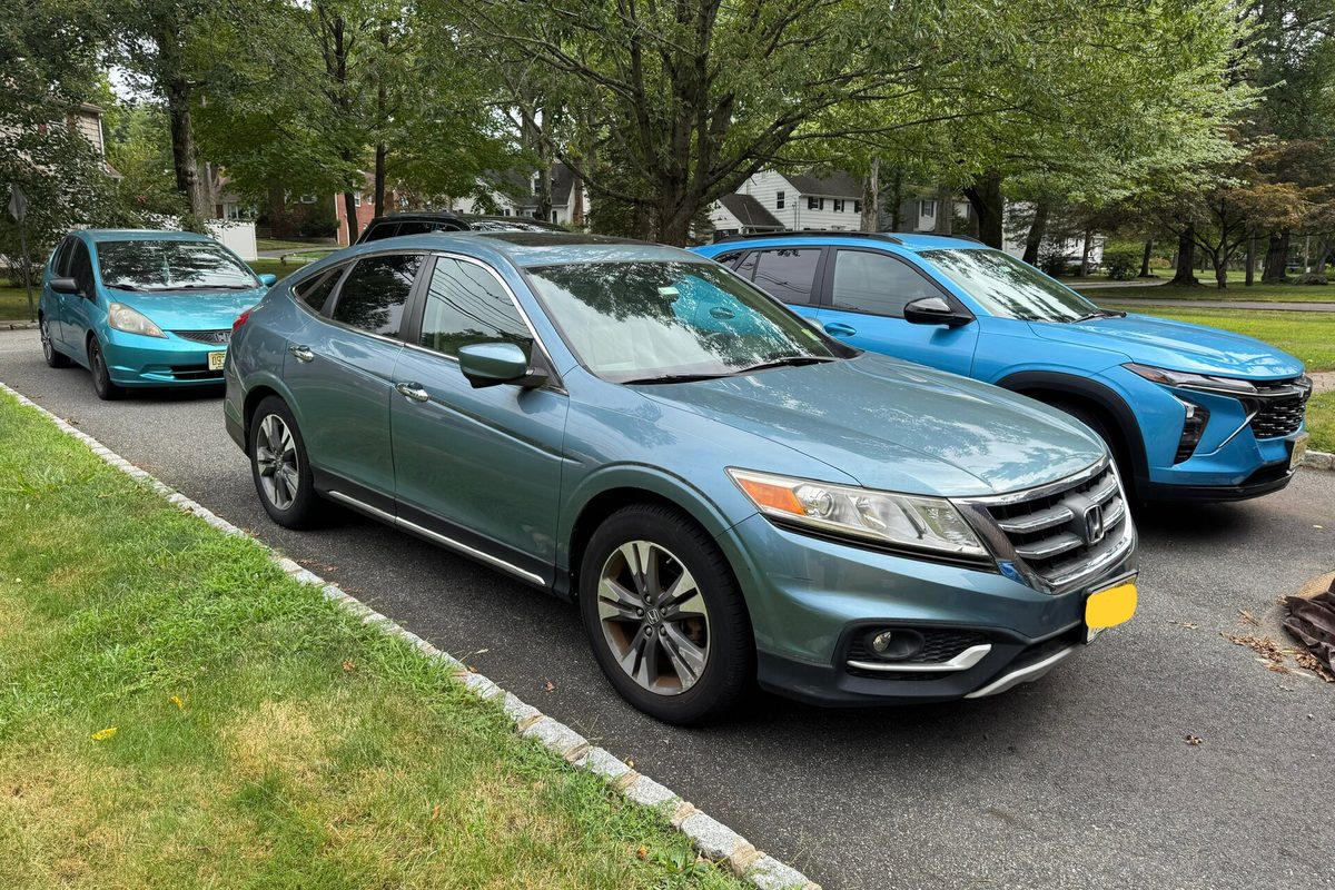 A blue-gray 2014 Honda Crosstour parked in a home driveway beside a lawn, with a teal hatchback and a blue SUV behind it