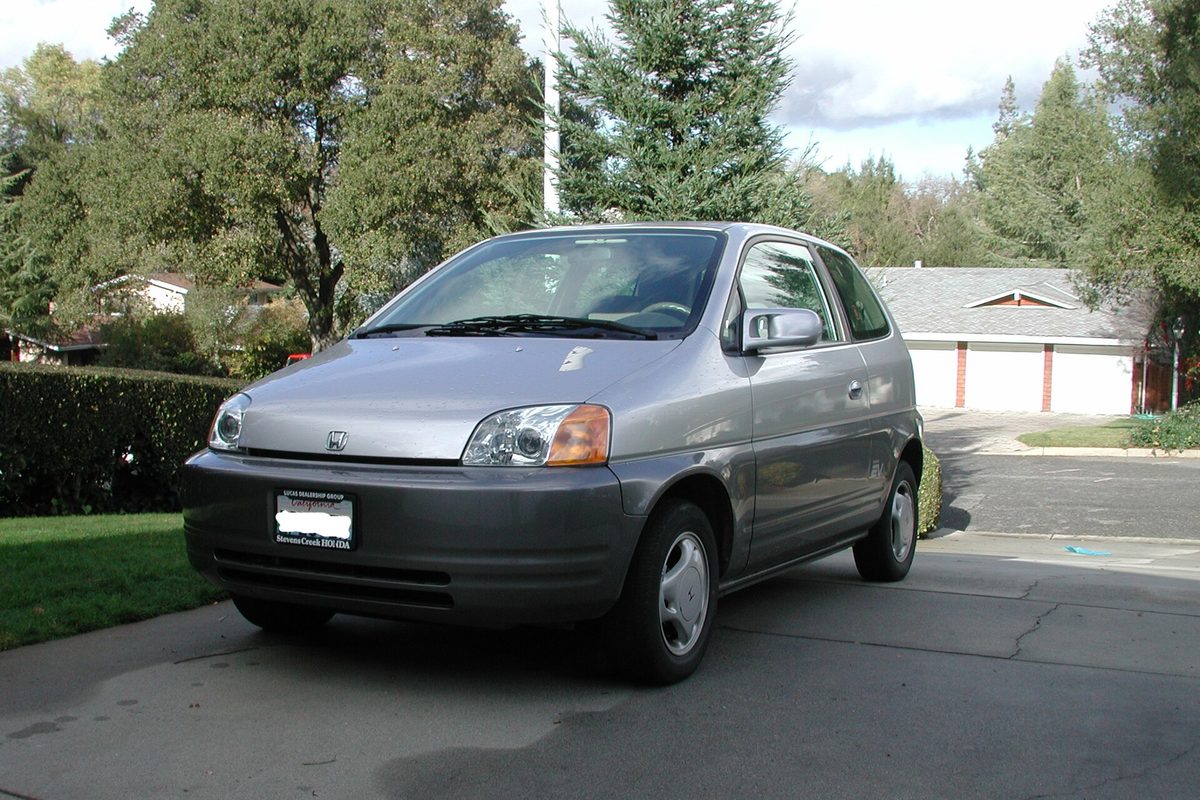 A silver Honda EV Plus three-door hatchback parked in a driveway, with a hedge, tall trees and a garage behind it