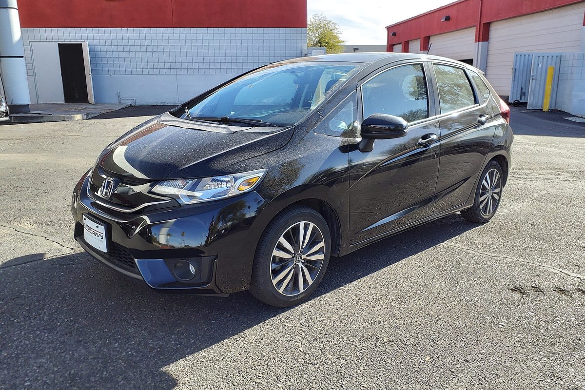 A black 2017 Honda Fit hatchback parked on sunny asphalt in front of a white and red industrial building