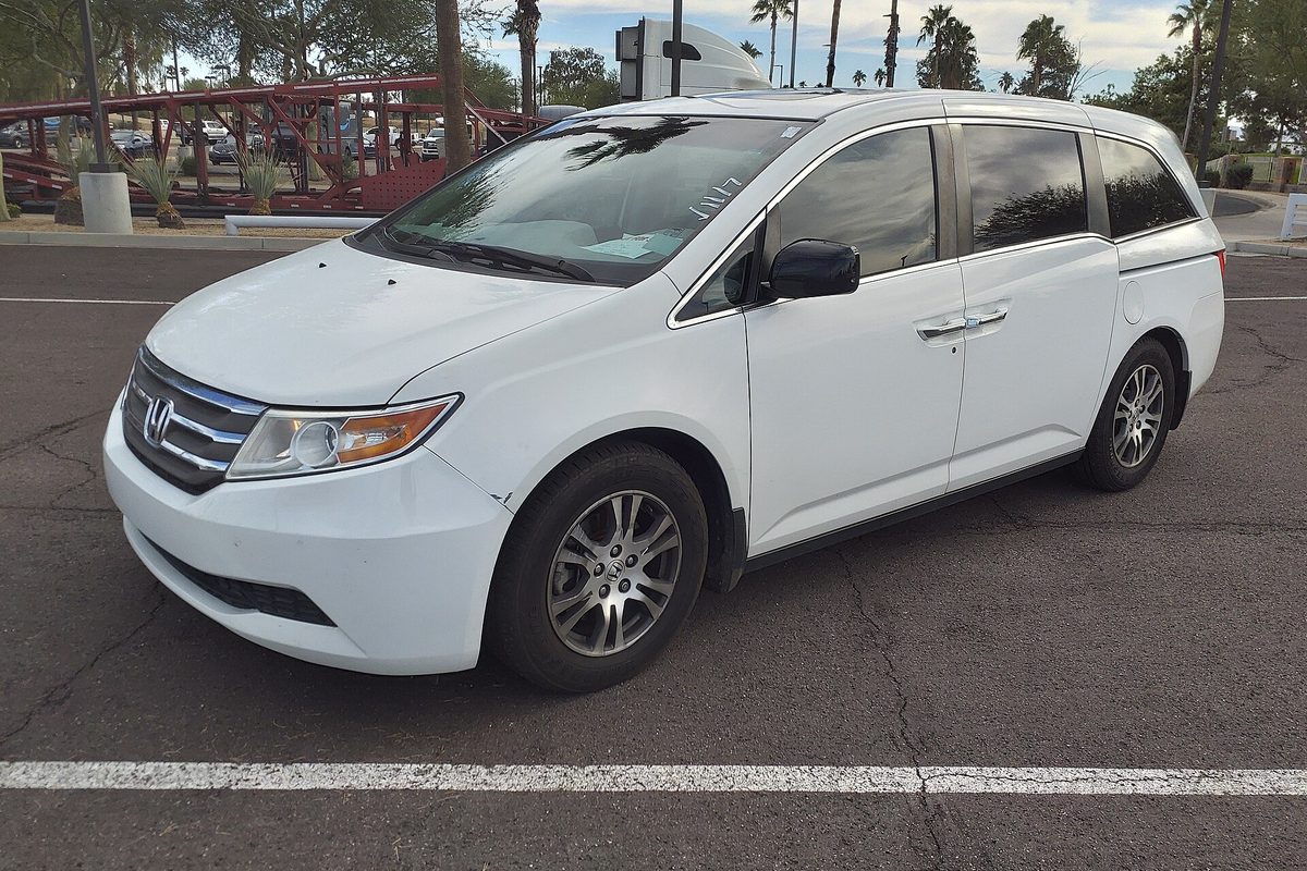 A white 2011 Honda Odyssey EX-L minivan parked in a sunny lot, palm trees and a red steel structure behind it