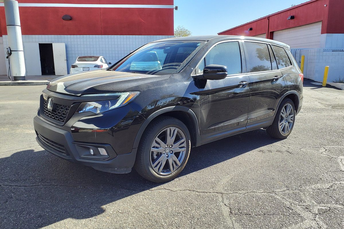 A black 2019 Honda Passport EX-L on cracked asphalt beside a red and white garage building