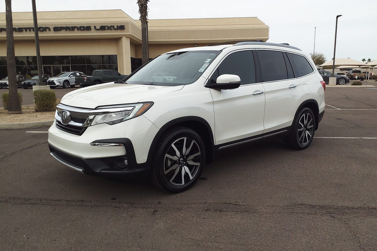 A white 2019 Honda Pilot Touring on a dealership lot under a grey sky, with other cars parked behind it