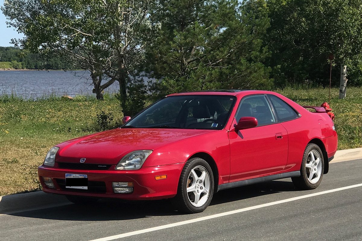 A red 2001 Honda Prelude coupe with a rear spoiler, parked at the roadside with a lake and trees behind it
